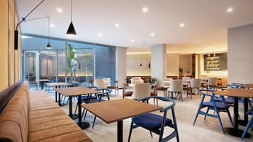 Un restaurant moderne avec des tables en bois et des chaises bleues, de grandes fenêtres et un espace détente avec un banc.