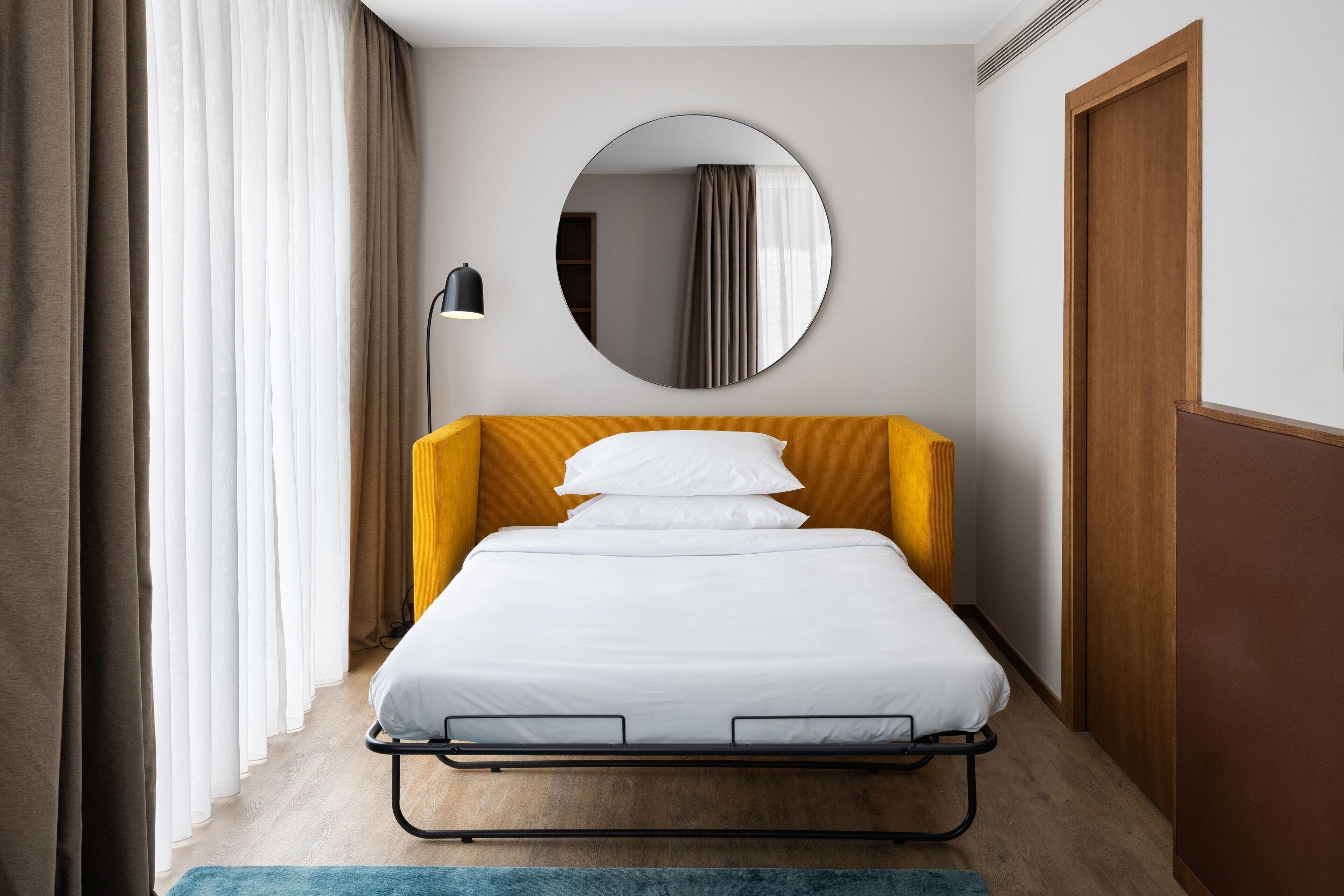Une chambre d’hôtel moderne avec un lit, un miroir rond et une porte en bois.