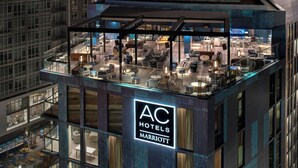 Un hôtel à plusieurs étages doté d’une façade en verre, d’un bar sur le toit et d’une enseigne bien visible indiquant « AC Hotels Marriott ».