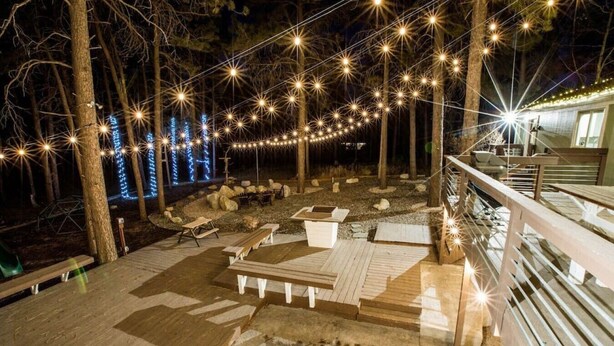 An outdoor deck with wooden benches and picnic tables, surrounded by trees and string lights. - THE FAMILY FOREST · ★3 Acres Of Forest★Lighted Yard★Chef Kitchen★ (Colorado Springs)