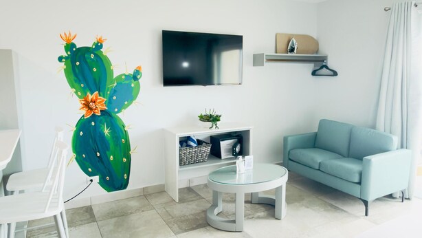 A modern hotel room with a flat-screen TV, a round coffee table, a light blue sofa, a white desk, and a cactus decoration. - Bushiri's Long Life Studio w/Kingbed & Balcony #5 (Oranjestad)