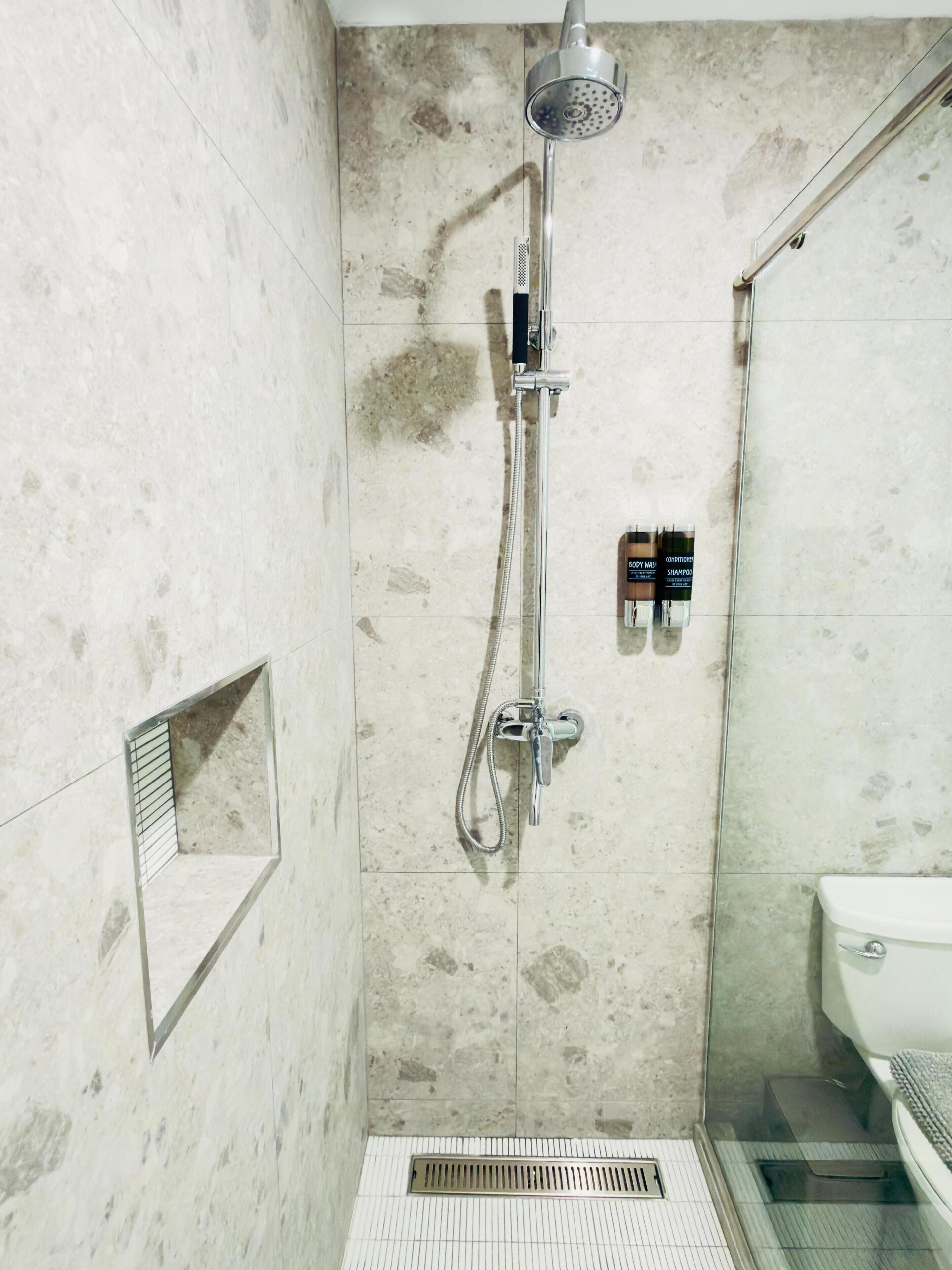 Bruser med håndbruser, brusearmatur og fastmonteret brusehoved. En lille hylde med toiletartikler og en gulvventil.