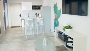 A modern kitchen with white cabinets, a microwave, and a sink. There are two white bar stools and a glass table in the foreground. A cactus painting decorates the wall. - Bushiri's Long Life Studio w/Kingbed & Balcony #5 (Oranjestad)