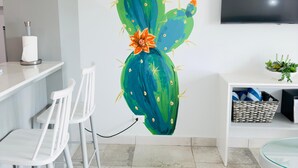 A modern kitchen with white chairs, a flat-screen TV, and a cactus wall art piece. - Bushiri's Long Life Studio w/Kingbed & Balcony #5 (Oranjestad)