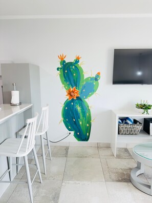 Une cuisine moderne avec des chaises blanches, un tĂ©lĂ©viseur Ă Ă©cran plat et une Ćuvre dâart murale reprĂ©sentant un cactus.
