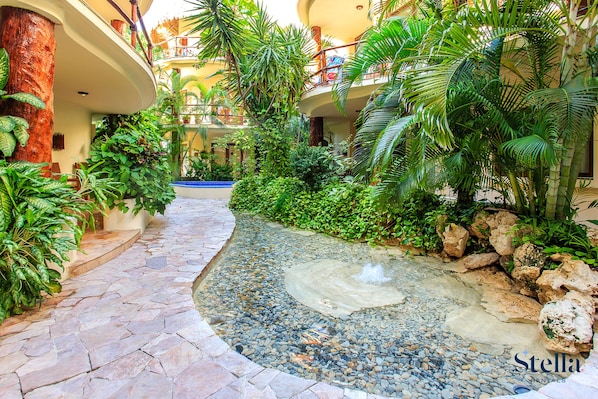 A tropical garden with a pebble-lined pathway, lush greenery, and a water feature.