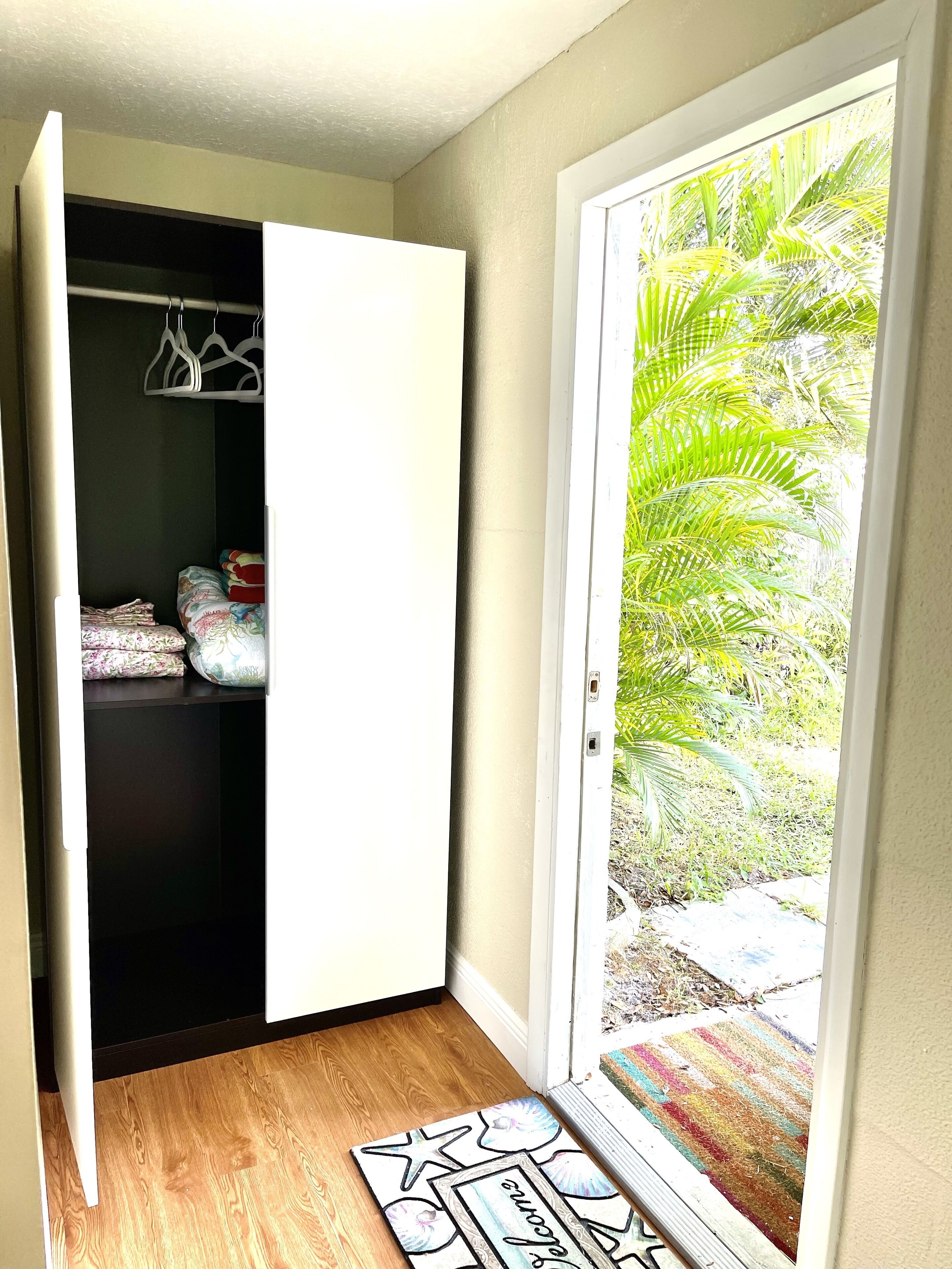 A room with a partially open closet, a wooden floor, and a door leading to a garden with palm trees.