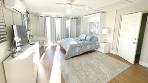 A bedroom with a bed, bedside tables, a desk, an air conditioner, a ceiling fan, and a door.