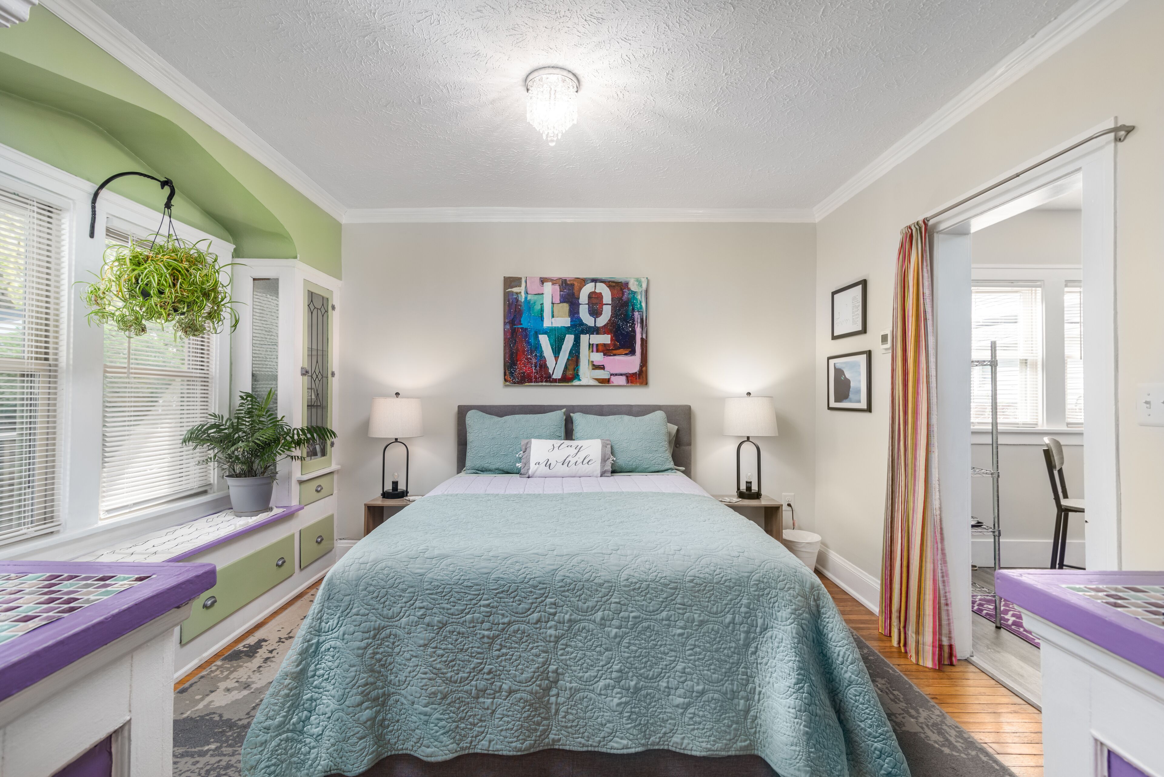 A bedroom with a bed, two bedside lamps, a window with blinds, a potted plant, a colorful artwork on the wall, and a striped shower curtain.