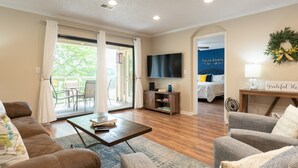 Ein geräumiges Wohnzimmer mit einer Couch, einem Sessel, einem Couchtisch und einem Fernseher. Ein Schiebeglastür führt zu einem Balkon mit Gartenmöbeln.
