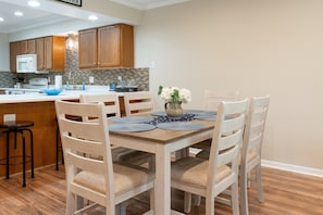 A dining area with a wooden table, four chairs, a vase with flowers, and a kitchen in the background.