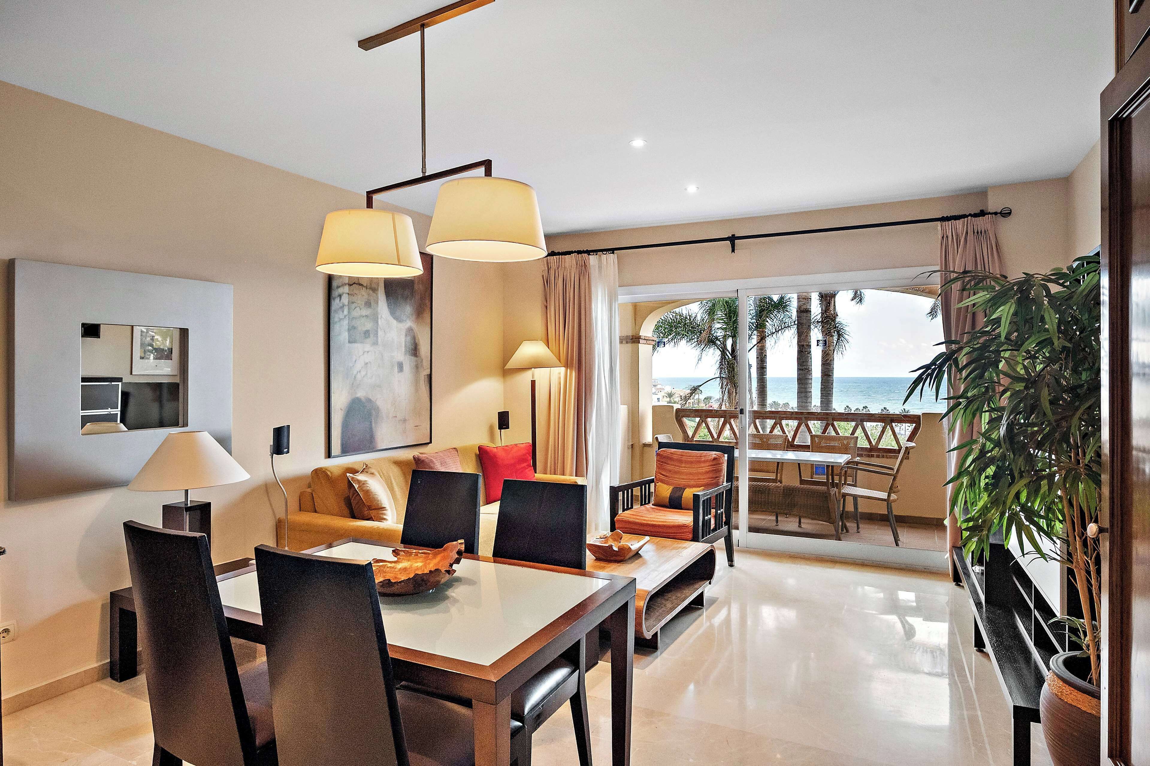 Ein modernes Hotelzimmer mit Essbereich, einer Couch, einem großen Fenster mit Blick auf Palmen und das Meer sowie einer Topfpflanze.