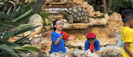 A group of children playing with toy guns in an outdoor setting with rocky terrain and a sign reading "WYNDHAM COSTA".