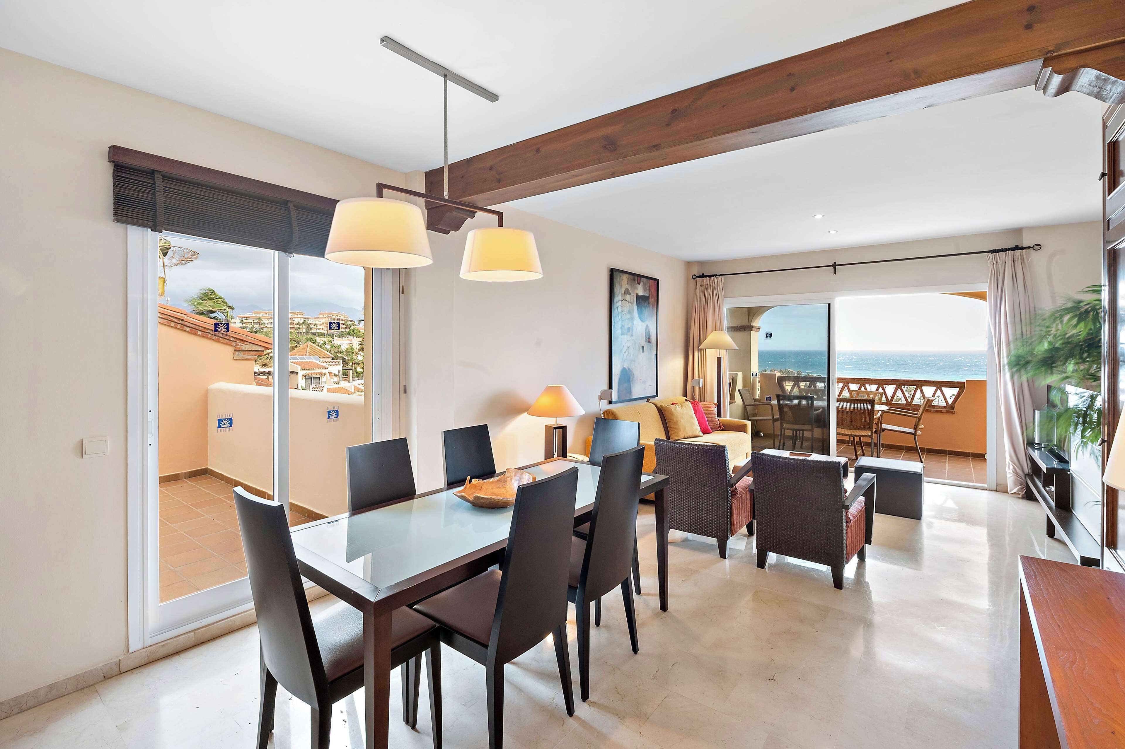 Ein geräumiges Wohnzimmer mit Esstisch, Stühlen und einer Couch. Ein Balkon bietet Blick auf das Meer.