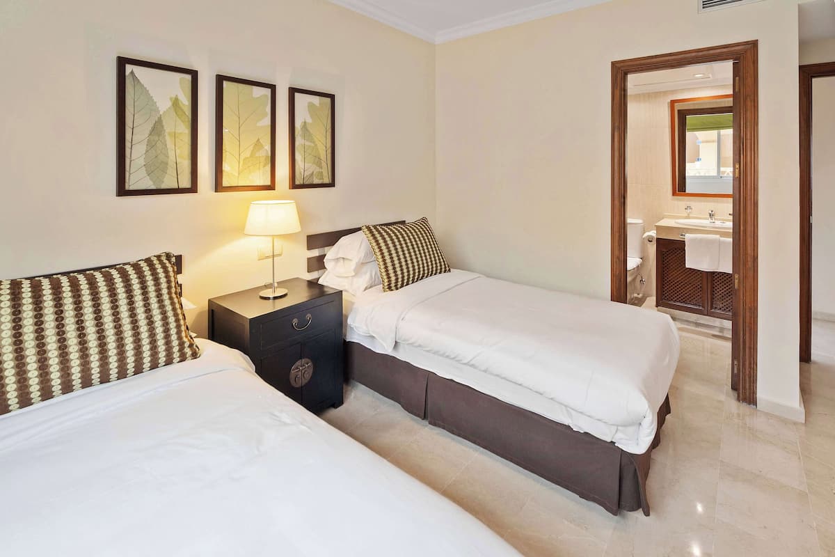 A hotel room with two beds, a nightstand with a lamp, and a bathroom visible through an open door.