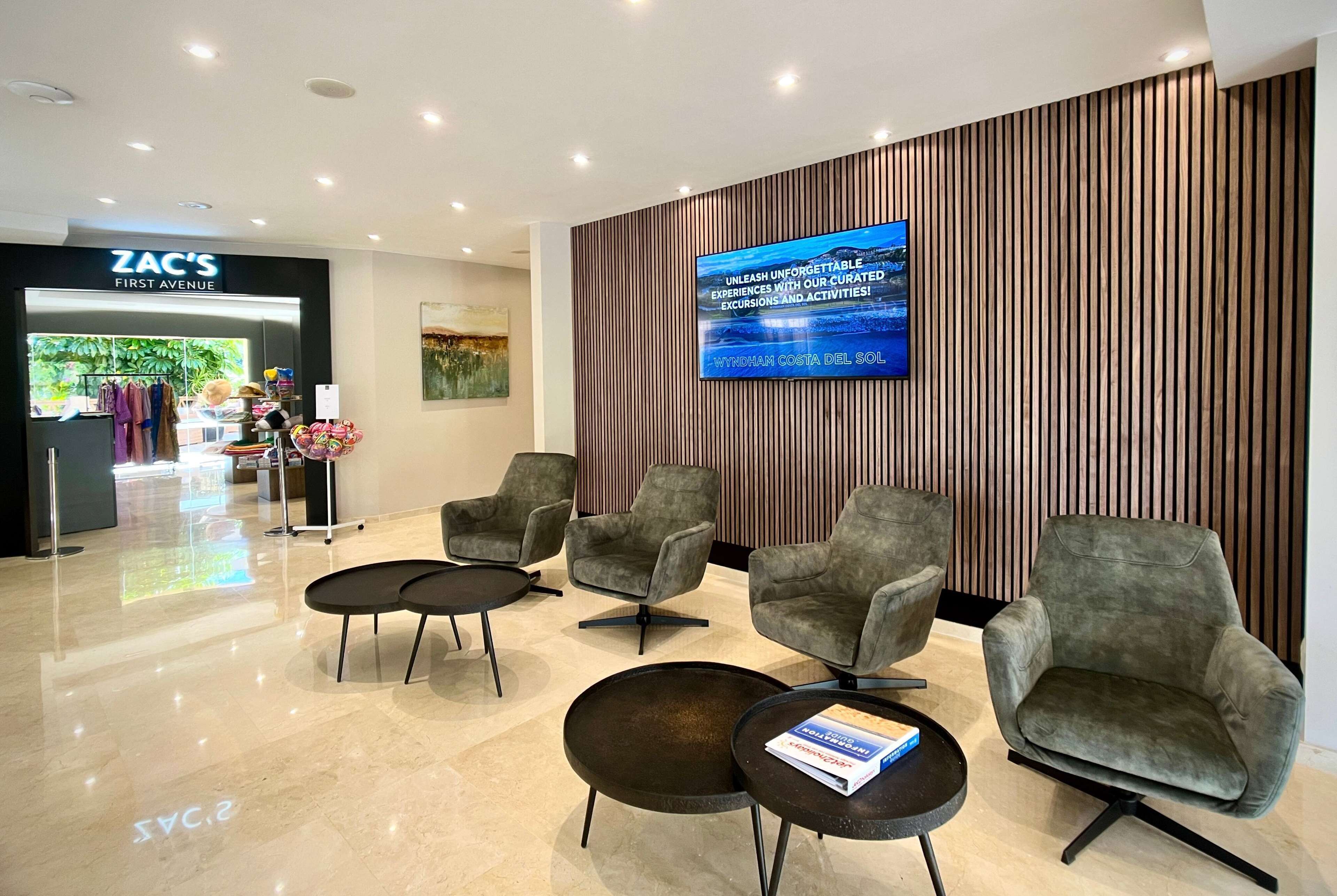 Eine moderne Lobby mit grau gepolsterten Sesseln, einem großen Flachbildfernseher an der Wand und einem Zara Geschäft im Hintergrund.
