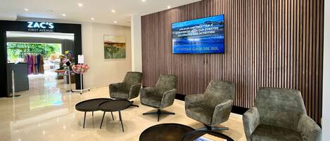 A modern lobby with grey upholstered chairs, a large flat-screen TV on the wall, and a Zara store in the background.