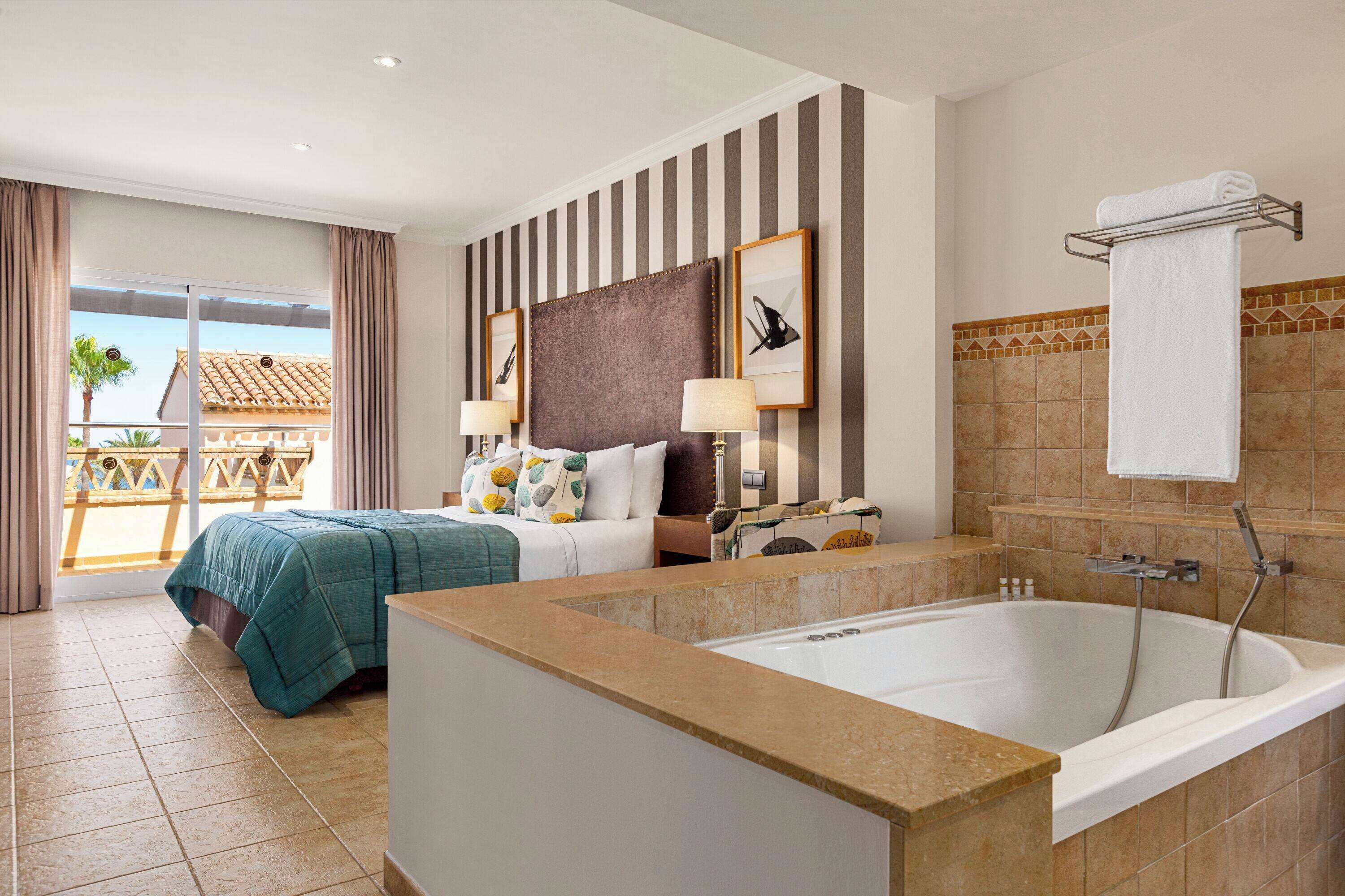 A hotel room with a large bed, a bathtub, a balcony with a view, and striped walls.