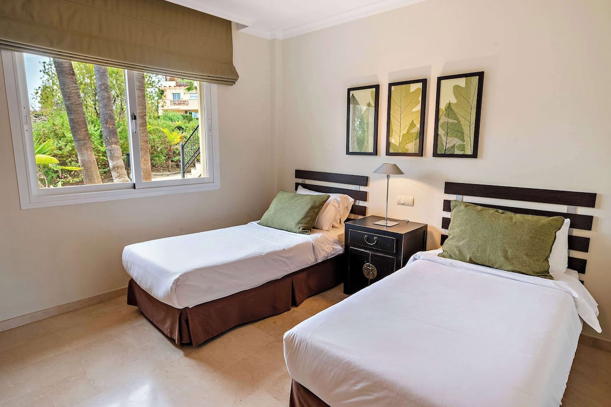 A hotel room with two beds, a window with a view of trees, and three framed leaf pictures on the wall.