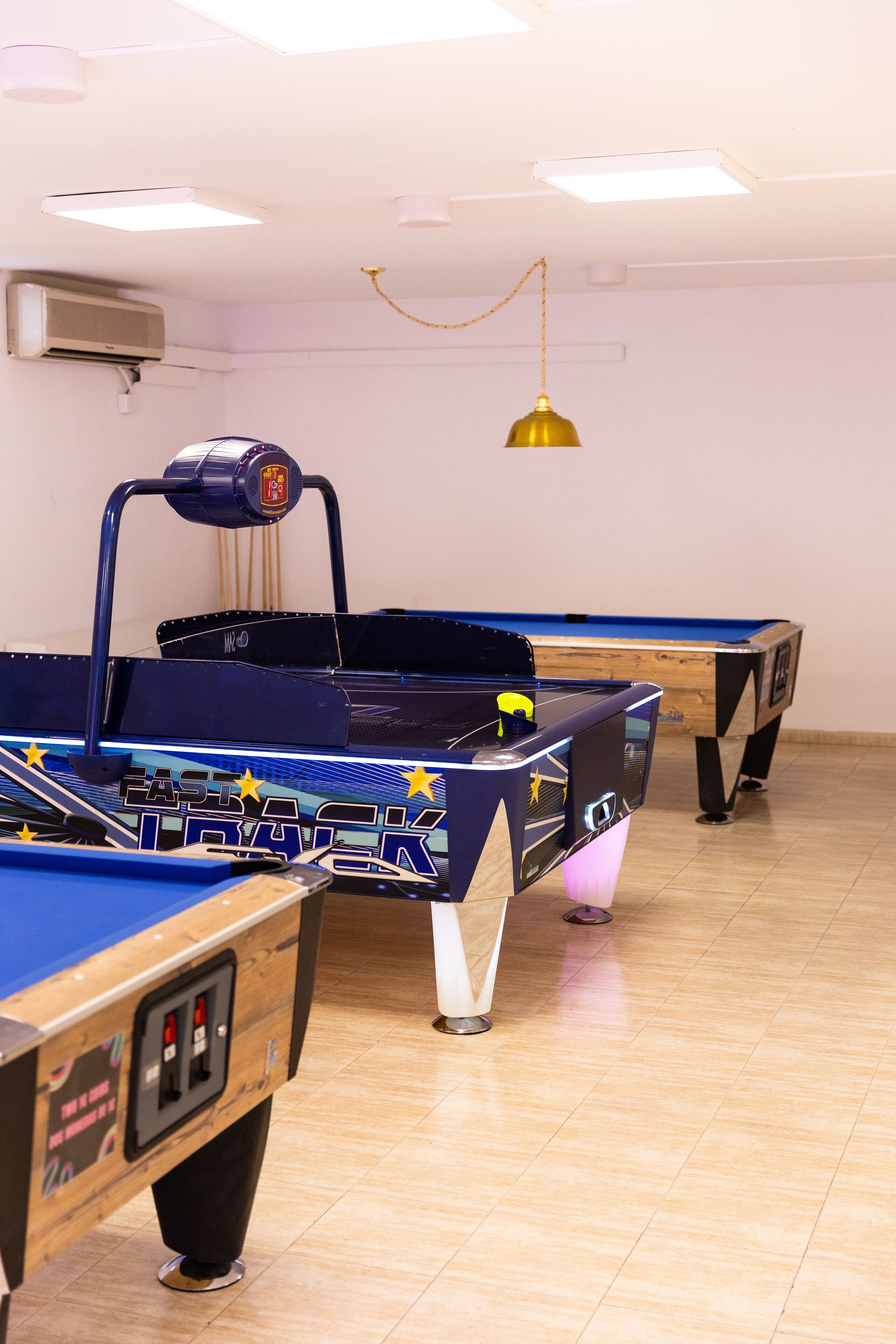 Uma área de jogos com várias mesas de bilhar, iluminação pendente e um aparelho de ar condicionado na parede.