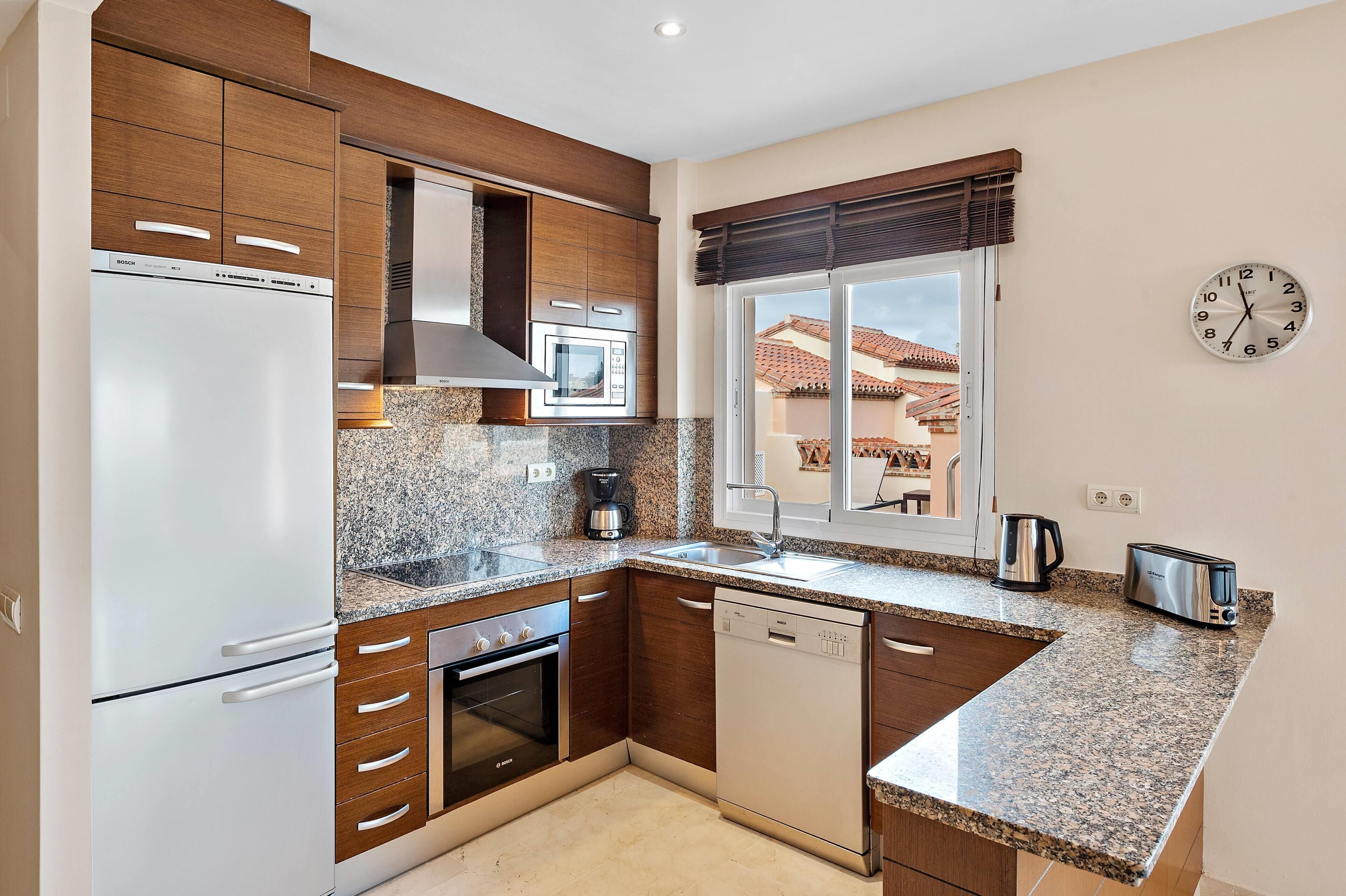 Une cuisine moderne équipée d’un réfrigérateur, d’un four, d’un lave-vaisselle et d’un four à micro-ondes de la marque Bosch. Une fenêtre offre une vue sur une maison, et une horloge est accrochée au mur.