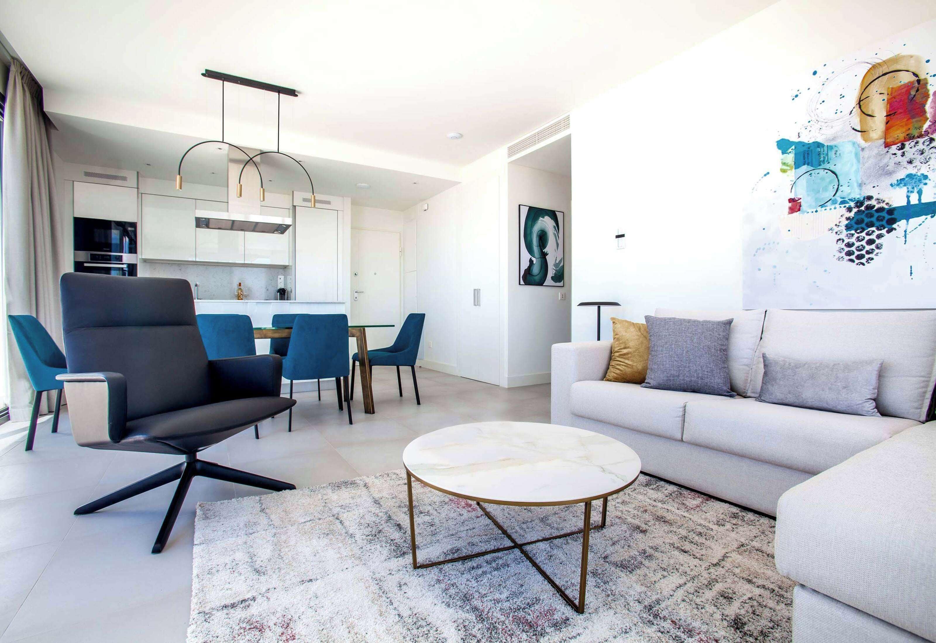 Un salon moderne avec une table basse ronde, un canapé et un coin repas comprenant des chaises, avec une cuisine en arrière-plan.