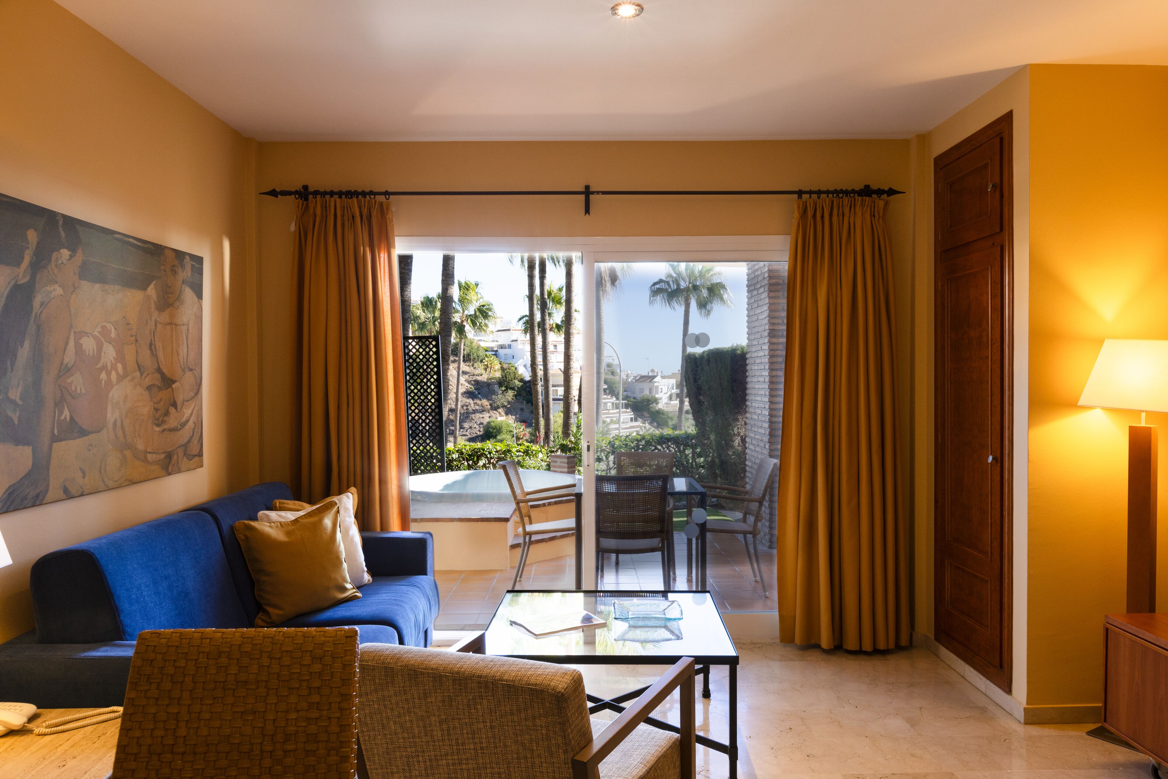 A hotel room with a blue sofa, a wicker chair, a glass table, a painting on the wall, and a view of palm trees and a pool outside.