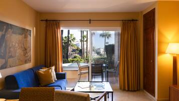 Une chambre d’hôtel avec un canapé bleu, un fauteuil en osier, une table en verre, un tableau au mur et une vue sur des palmiers et une piscine à l’extérieur.