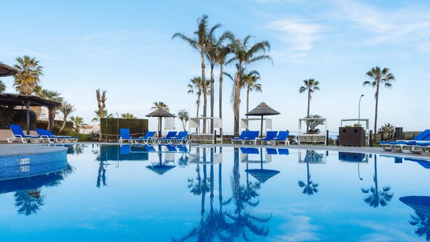 Zona de piscina con sillas reclinables azules, palmeras y cielo azul despejado.