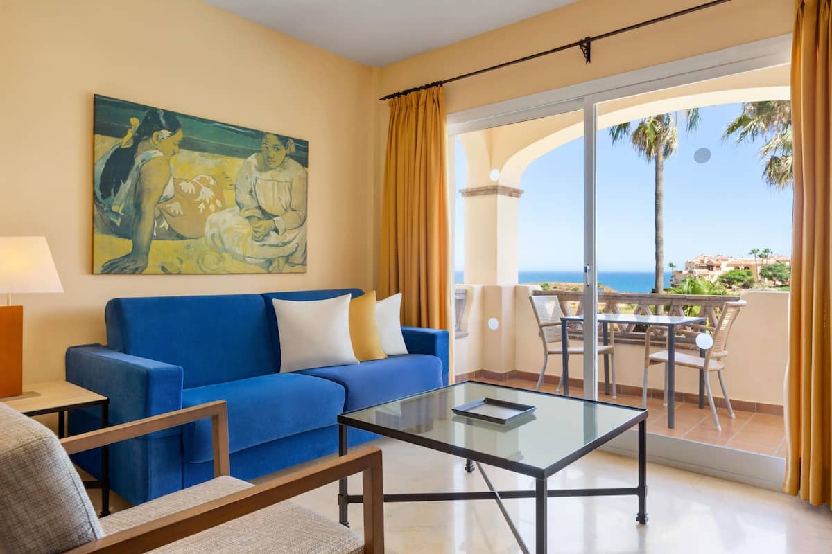 A room with a blue sofa, a glass coffee table, a painting on the wall, and a balcony with a view of the sea.