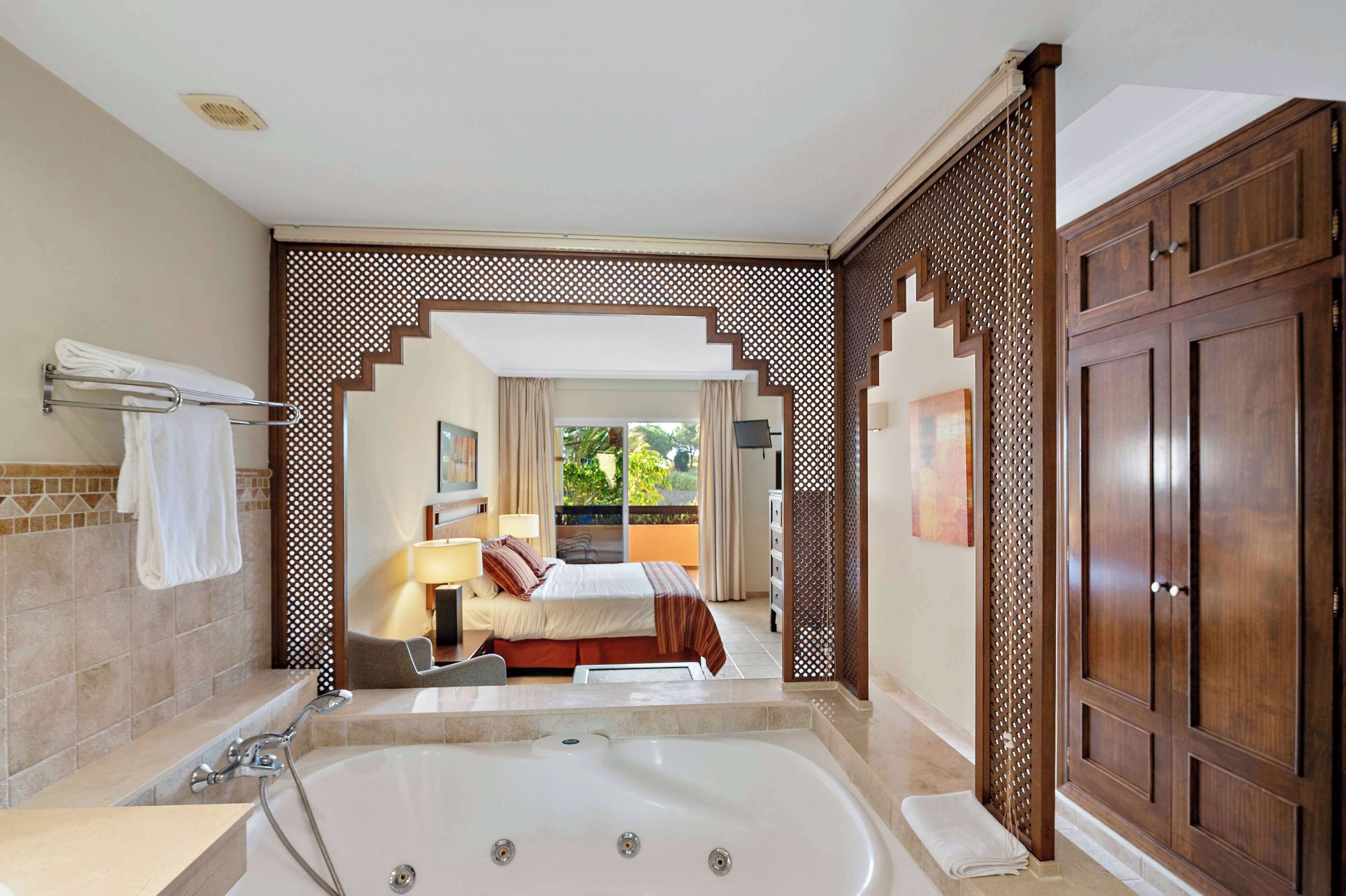 A modern bathroom with a large bathtub, a shower, and a toilet. There is a bed with a view of the outdoors.