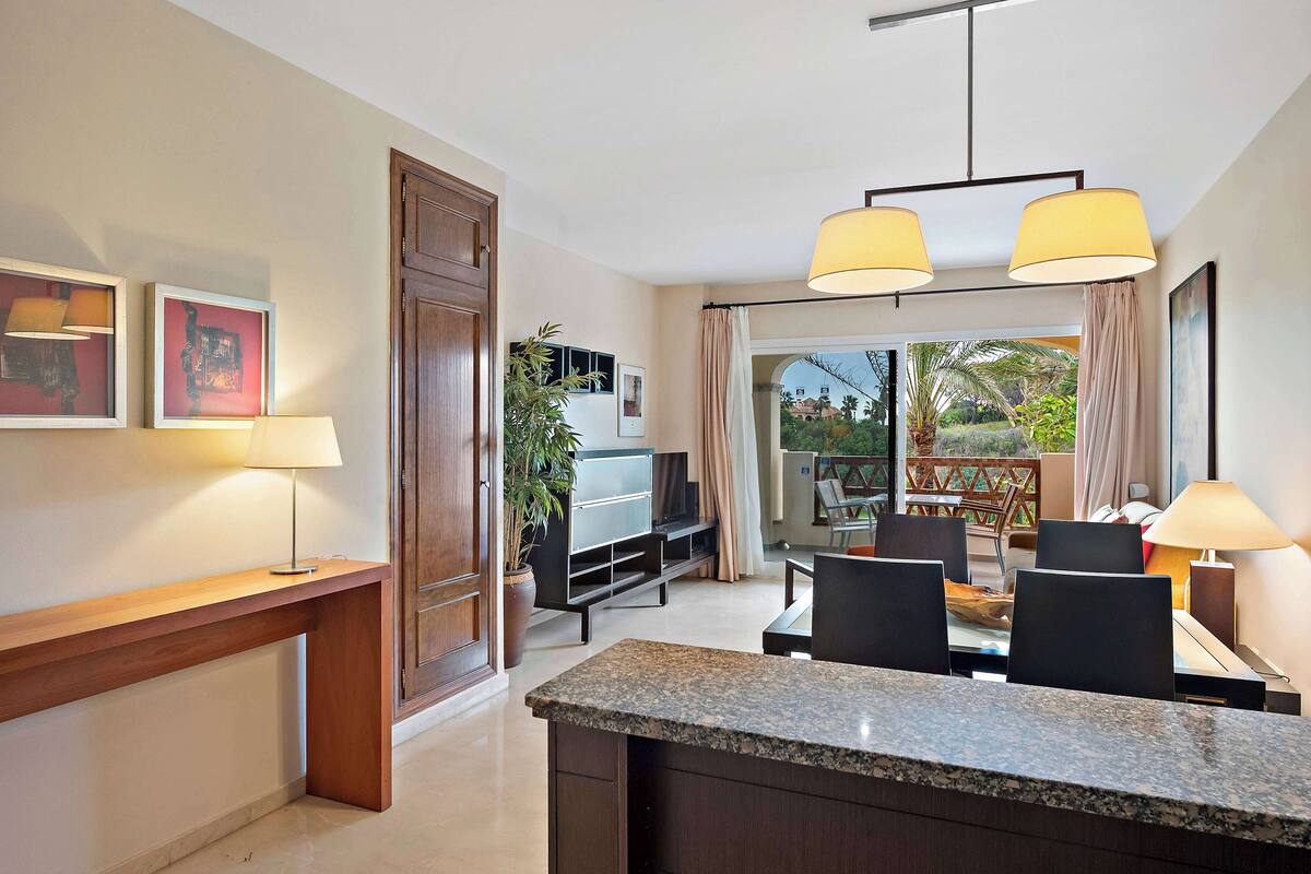 A modern hotel room with a dining area, a TV, a plant, and a balcony with a view.