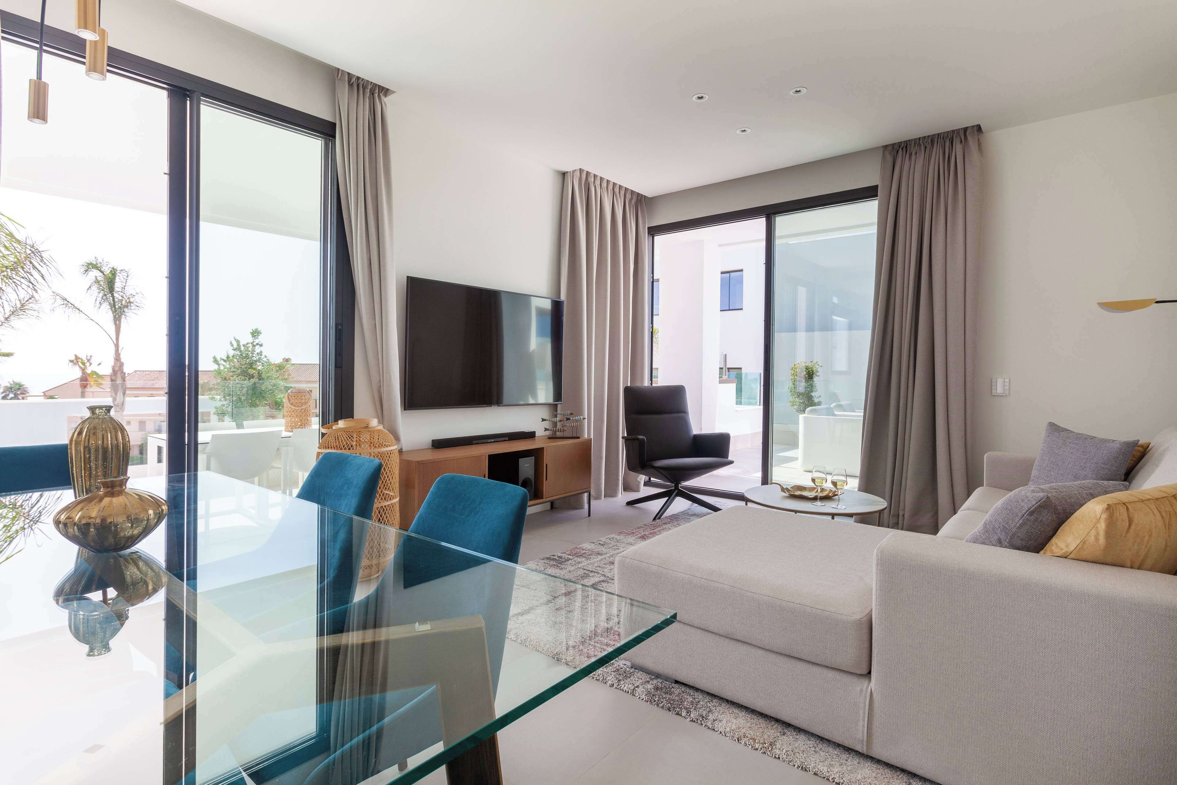 A modern living room with a glass dining table, a sofa, a TV, and a balcony with curtains.