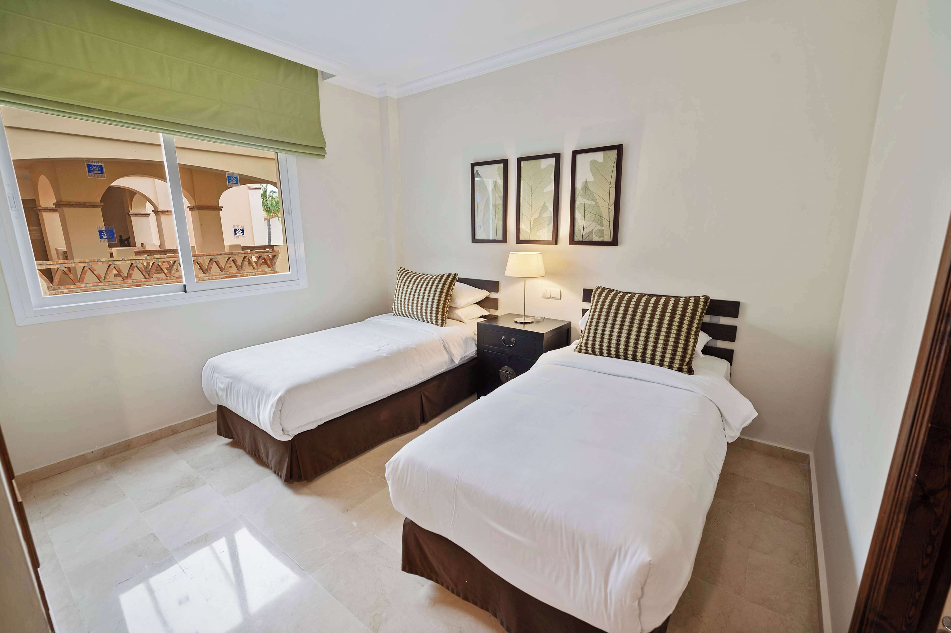 A hotel room with two beds, a window with a view of an outdoor area, and a wall with three framed pictures.