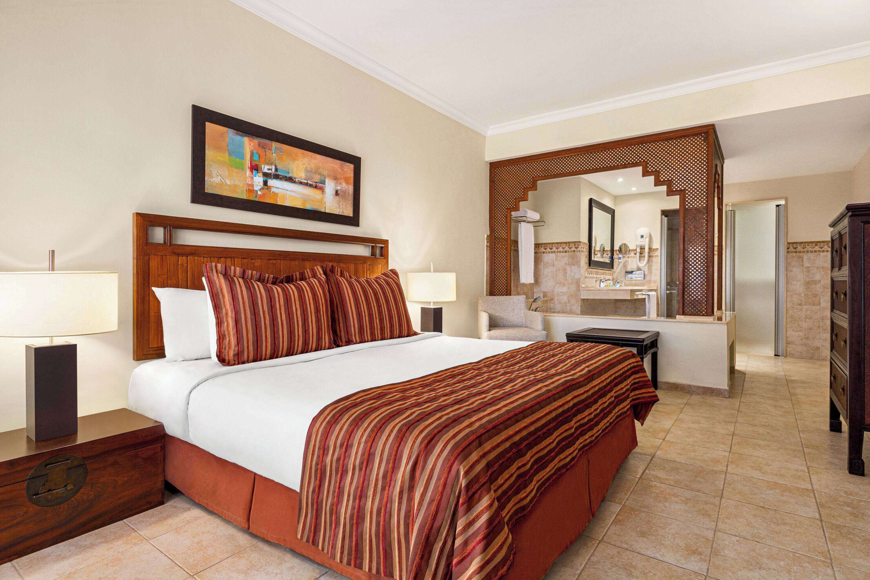A hotel room with a large bed, two bedside tables with lamps, a wooden headboard, a painting above the bed, a bathroom with a mirror, and a wardrobe.