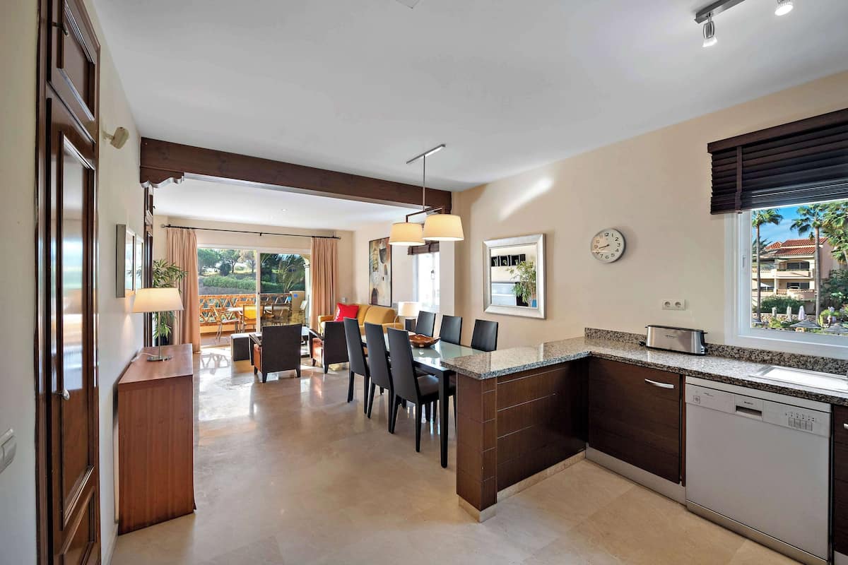 A modern kitchen with a built-in dishwasher, a dining area with a table and chairs, and a view of the outdoors through a window.