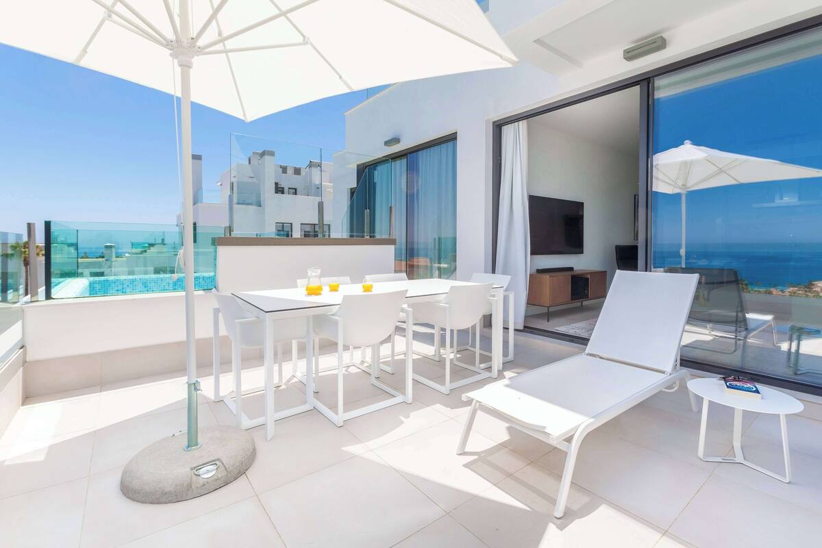 A modern outdoor balcony with a dining area, lounge chairs, and a view of the ocean.