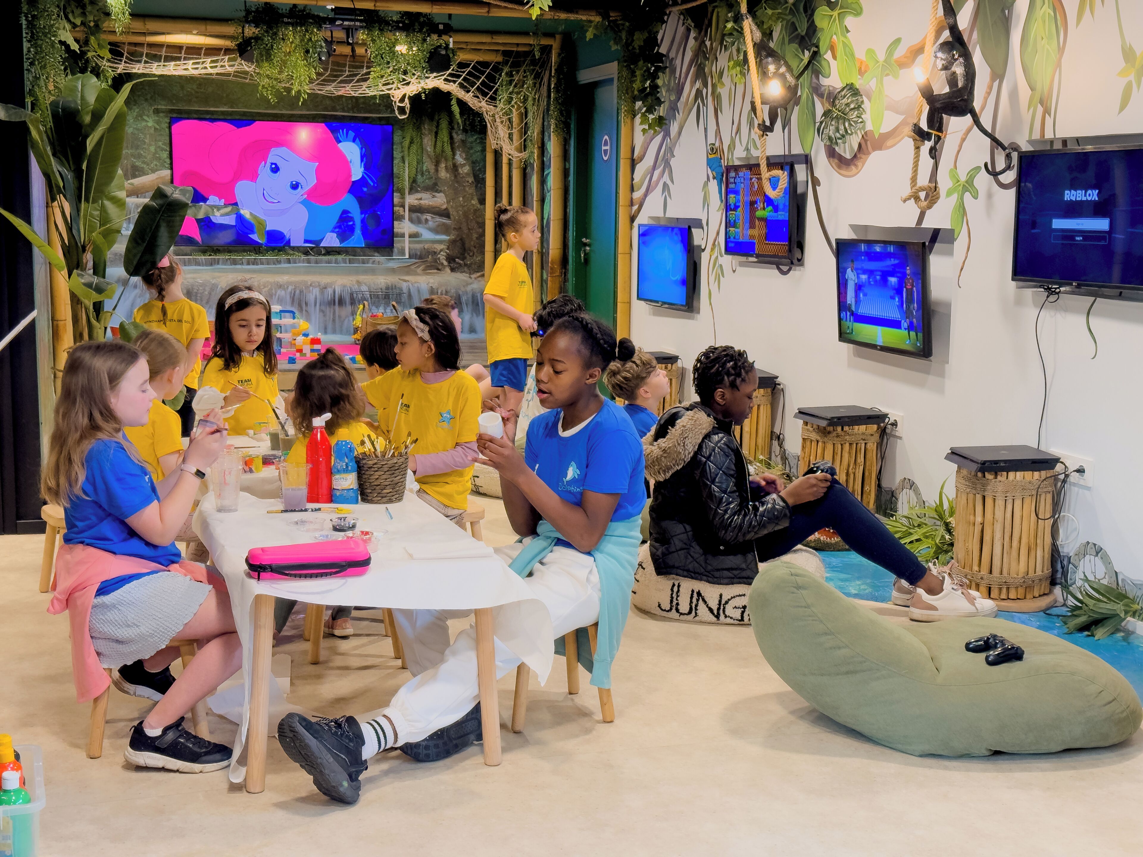A group of children engaged in an art activity in a room with a jungle theme, including a large TV screen and multiple smaller screens.