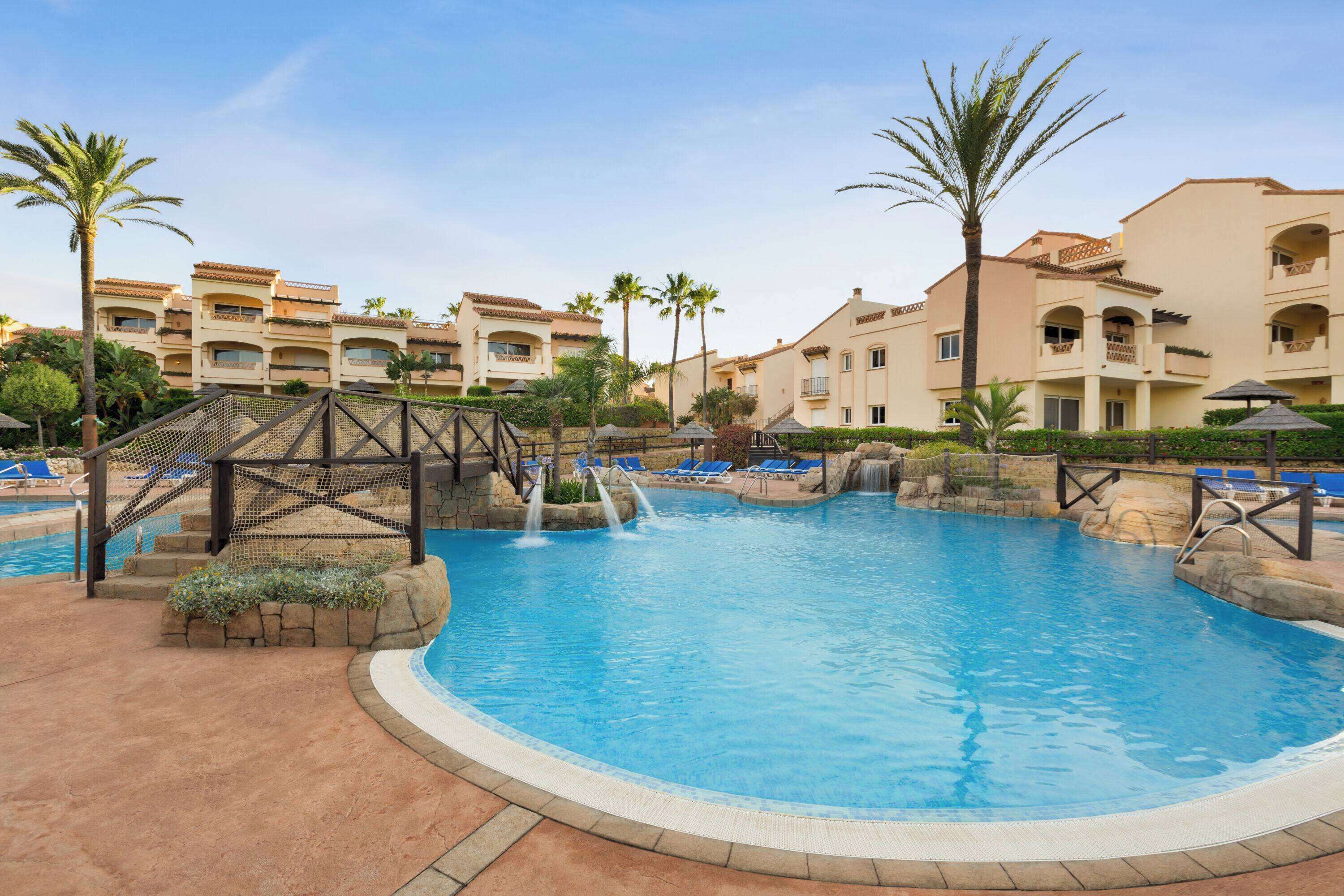 Una piscina con cascada, rodeada de palmeras y un edificio de varios pisos con balcones.