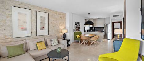 A modern living room with a sofa, coffee table, and a bright yellow armchair. There is a dining area with a table and chairs, and a kitchen in the background.