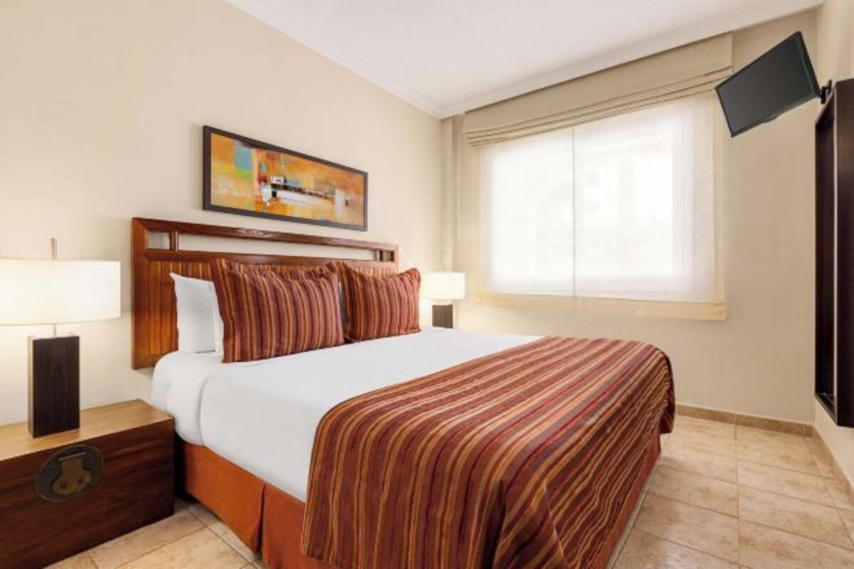 Una habitación de hotel con una cama grande, dos mesitas de noche, un televisor de pantalla plana montado en la pared y una ventana con cortinas.