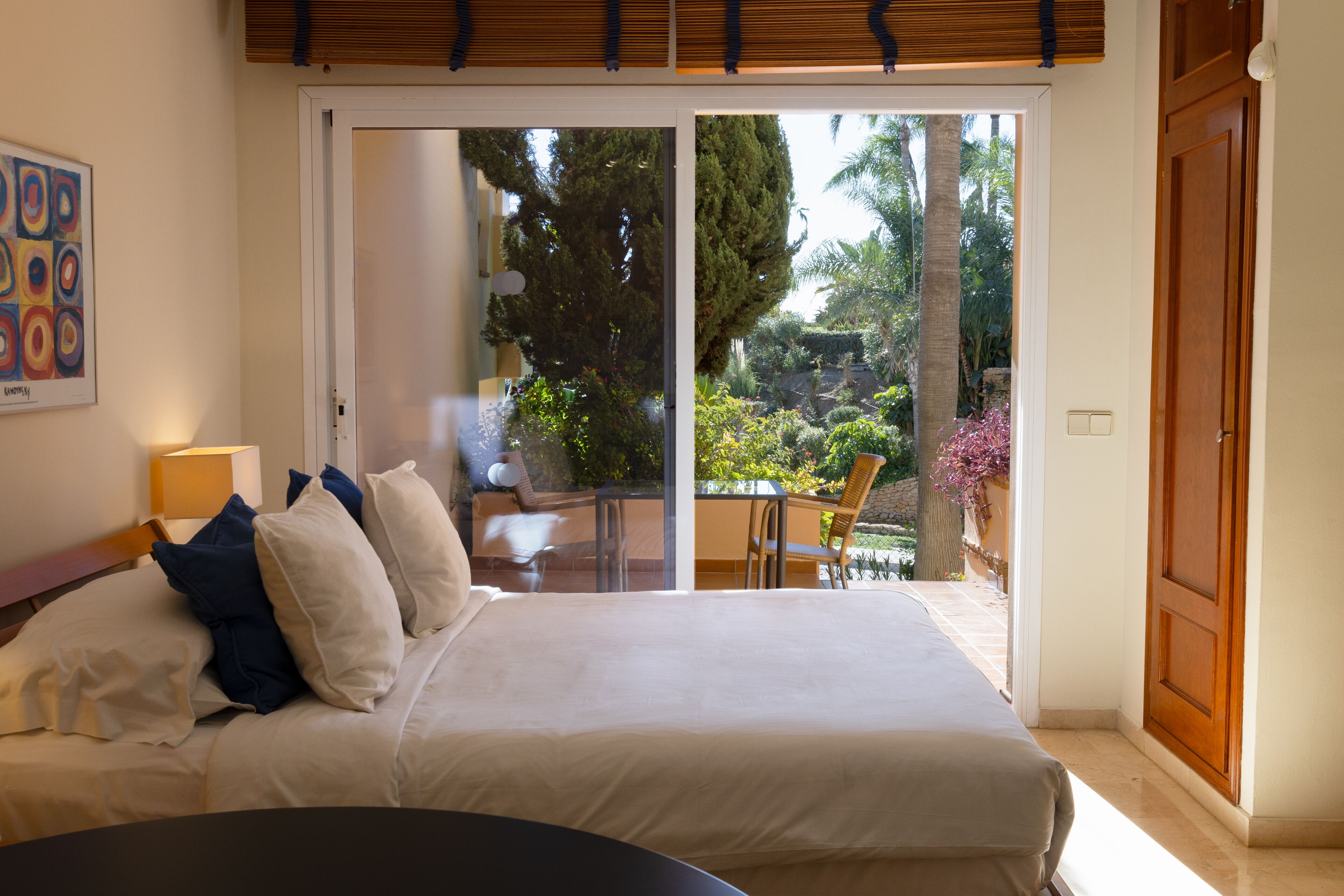 A bedroom with a large bed, a desk, and a chair. There is a sliding glass door leading to a balcony with plants and a table.