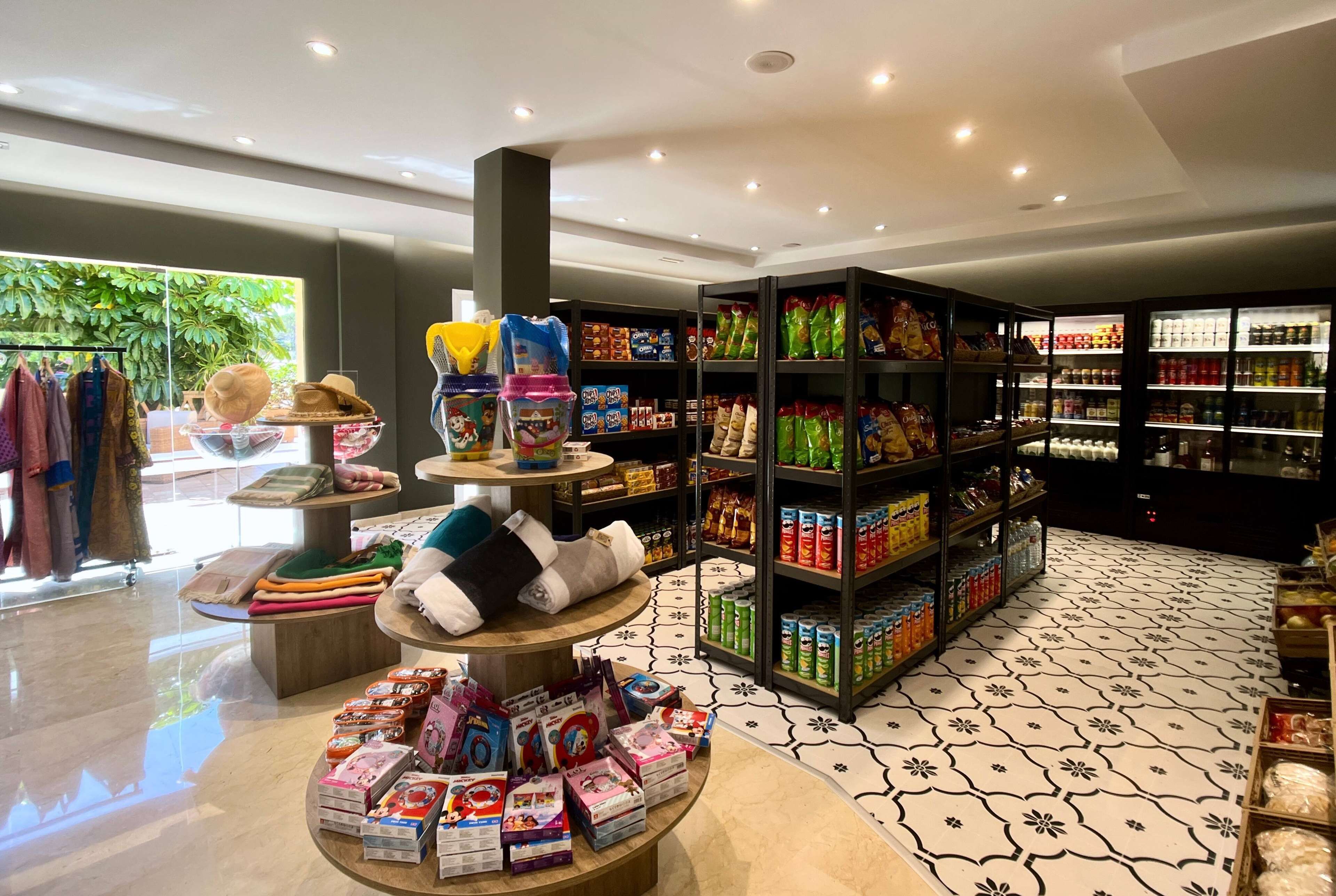 Un magasin bien éclairé, avec des étagères remplies d’en-cas et de produits emballés, un présentoir de chapeaux et de vêtements pliés, et un sol à motifs.