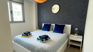 Une chambre avec un grand lit, une table de chevet, une commode, un luminaire suspendu, une fenêtre et un miroir au mur.
