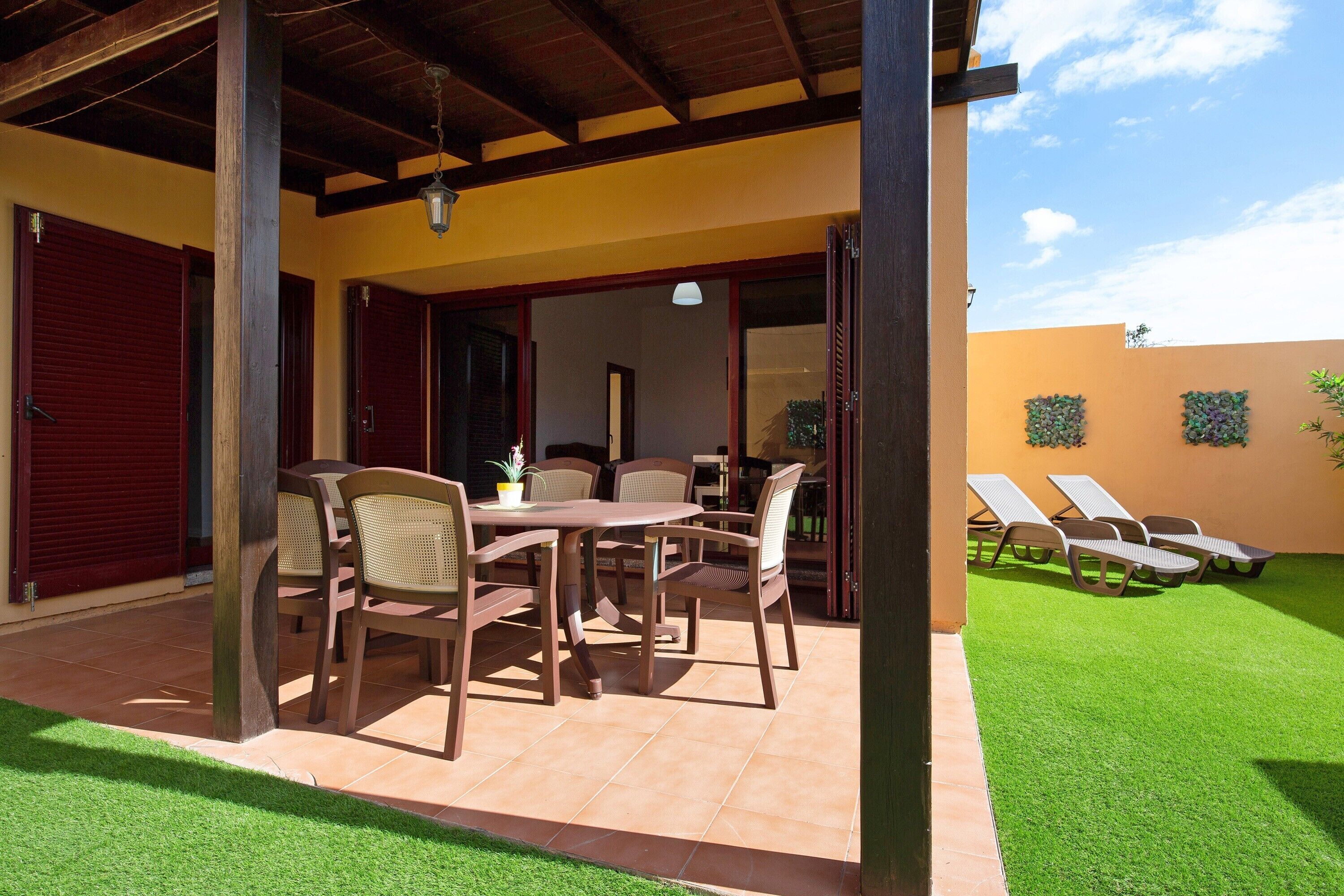 Une terrasse couverte avec une table et des chaises, une pelouse et un bâtiment avec un volet rouge.