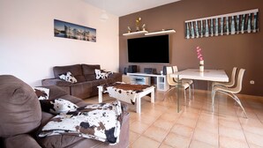Un salon moderne comprenant un canapé, une table basse, un téléviseur, une table à manger avec des chaises, et des œuvres d’art accrochées aux murs.