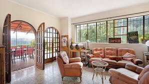 A spacious living room with leather furniture, a large window with a view of greenery, and a door leading to an outdoor area.