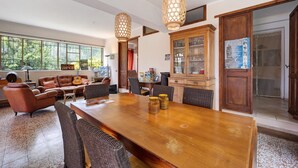 A spacious living room with a large wooden dining table, comfortable seating, and a view through large windows.