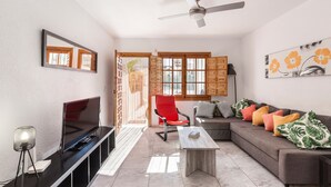Una sala de estar con televisor, una butaca roja, un sofá con cojines, un ventilador de techo y una ventana con contraventanas de madera.