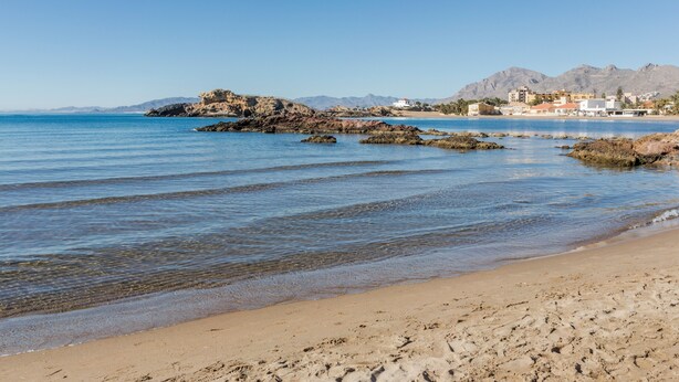 Una playa con agua cristalina, afloramientos rocosos y montañas a lo lejos.